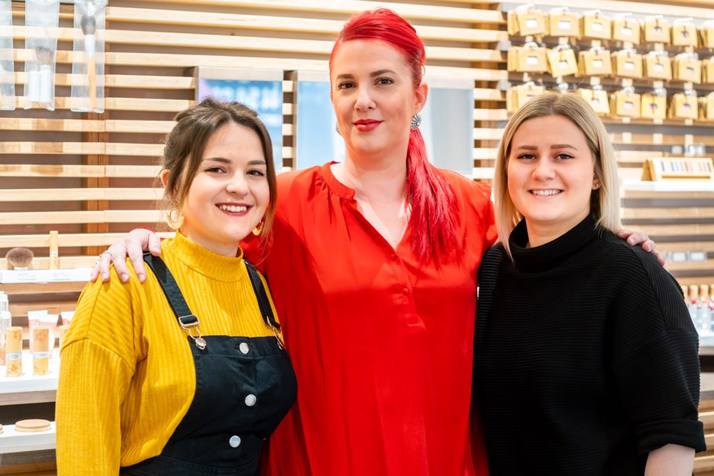Tre professioniste del salone sorridenti posano insieme abbracciate davanti a una parete a doghe di legno che espone prodotti cosmetici. La donna al centro ha i capelli rosso acceso e una maglia rossa.